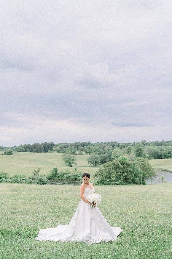 Bridal portrait of a bride holding a white bouquet in a strapless gown with long train, looking down in a grassy field by a pond under cloudy sky