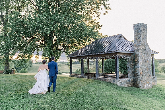 Couple portrait of bride in a strapless wedding dress and groom in a blue suit walking away under pavilion string lights at sunset