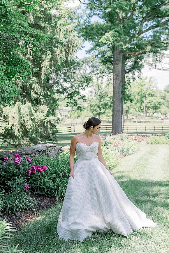 Bridal portrait of a bride in a strapless ball gown wedding dress, holding her skirt and looking down on a garden lawn by stone wall