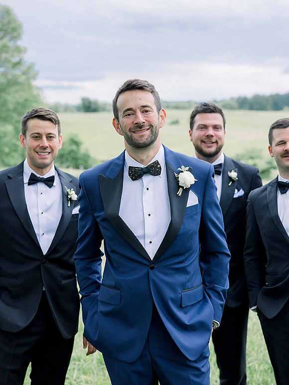 Groom portrait in a blue tuxedo with black lapels and boutonniere, hands in pockets, standing in a field with rolling hills under cloudy sky