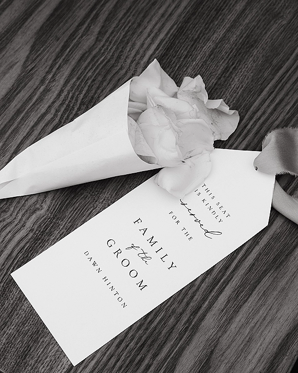 Wedding place card with calligraphy escort card lettering beside a paper flower petal cone tied with ribbon on a wood table flatlay