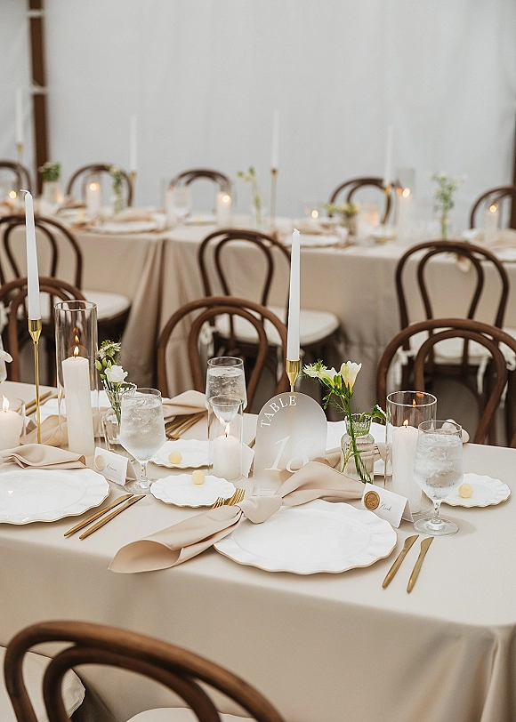 Reception tablescape with round wedding table setup featuring beige linens, white taper candles, bud vases, gold flatware, and bentwood chairs indoors