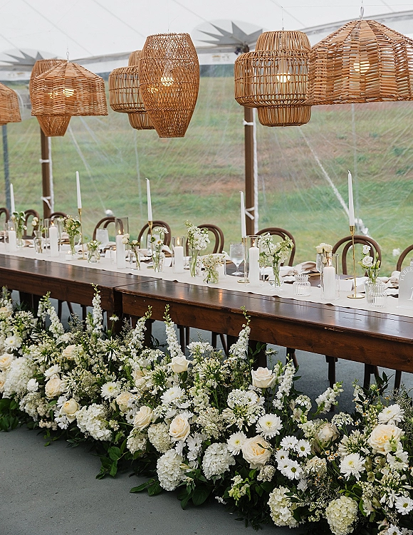 Reception tablescape with long banquet table decor, white runner, bud vases of roses and hydrangeas, greenery, candles under a clear tent