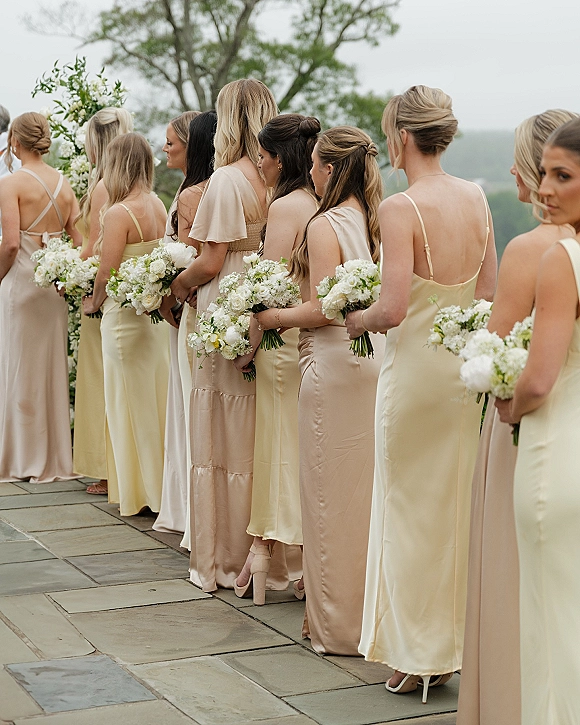 Bridesmaid lineup in champagne bridesmaid dresses holding white bouquets on a stone patio with trees, cloudy sky, and distant hills