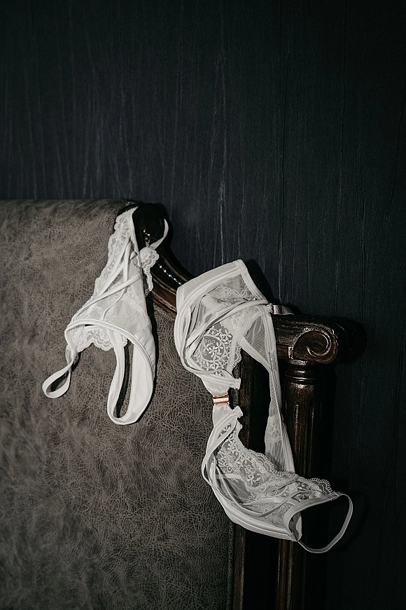 Bridal lingerie wedding night lingerie set in white lace with rose gold hardware draped over an upholstered armchair against a dark wall