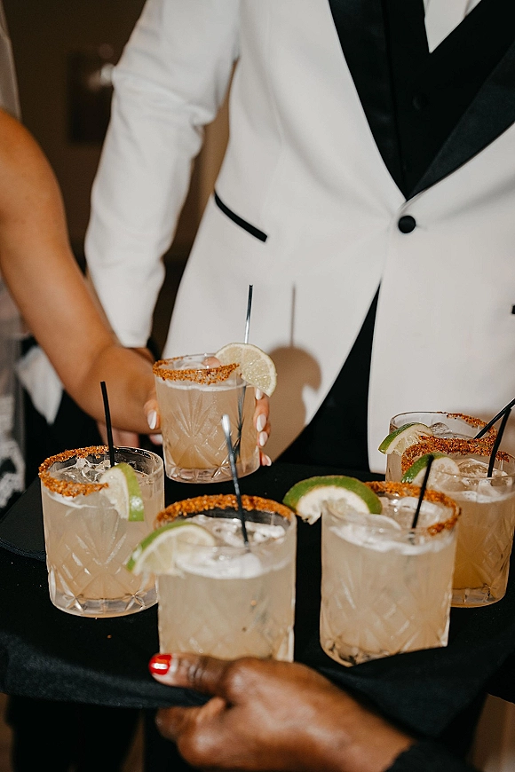 Wedding cocktails on a tray with ice, black straws, lime wedges, and chili salt rims, served by a waiter in a white jacket at reception