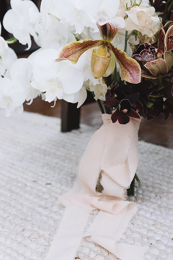 Wedding bouquet of cascading orchids tied with a blush ribbon in a vase on a textured rug with chair legs in the background