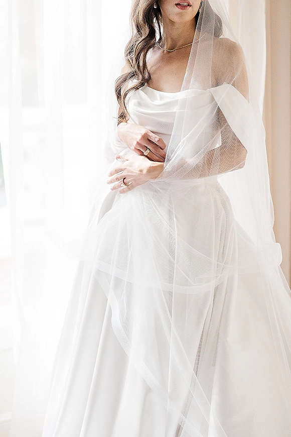Bridal portrait of a bride in veil wearing a strapless wedding dress, posing in soft window light beside curtains, drop earrings visible
