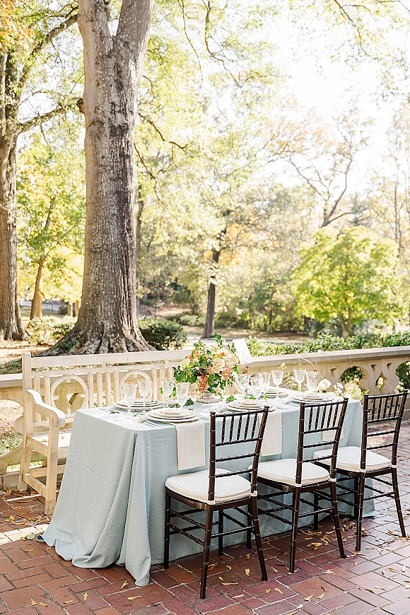 Reception tablescape with an outdoor reception table featuring light blue linens, floral centerpiece, china plates, and crystal glasses on a brick terrace