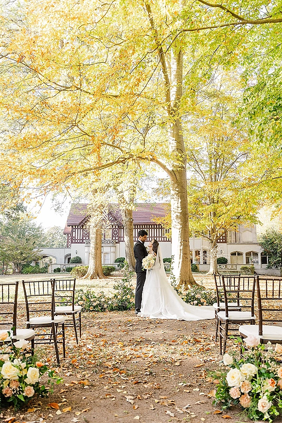 Couple portrait outdoor wedding portrait of bride and groom embracing, her bouquet and veil trailing by aisle flowers near a manor house in fall