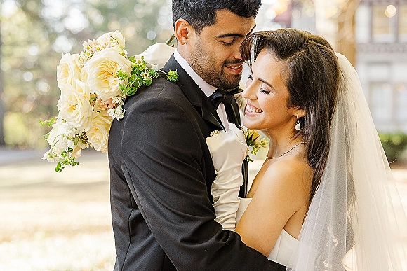 Couple portrait with bride and groom embrace, foreheads touching as she smiles, holding a white rose bouquet amid outdoor greenery