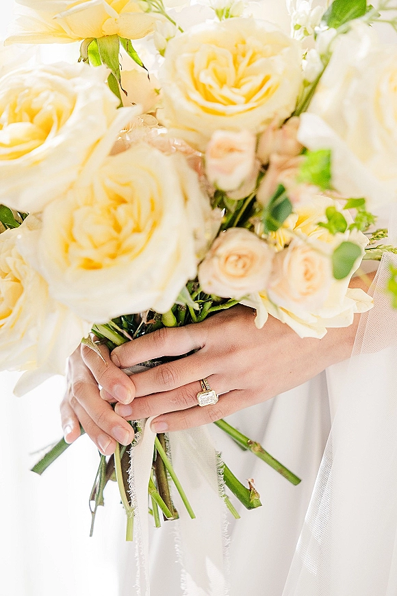 Bridal bouquet of cream roses and blush blooms with greenery, held in bridal hands showing an emerald cut engagement ring on a bright white backdrop