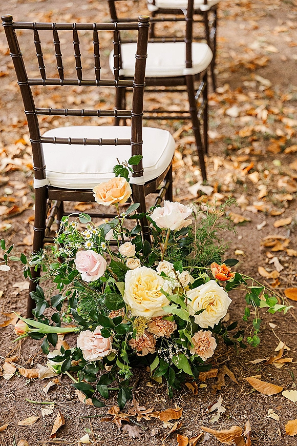 Ceremony aisle decor with an aisle floral arrangement of roses and greenery beside chiavari chairs on a dirt path with autumn leaves