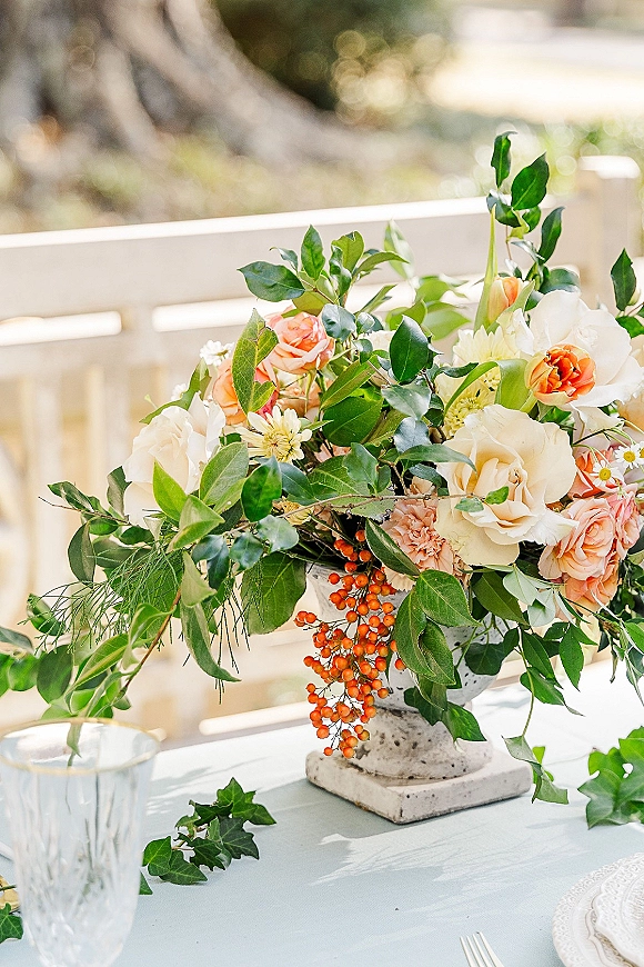Wedding centerpiece with a floral centerpiece of roses, ranunculus, and orange berries in a compote vase on a lace tablecloth outdoors