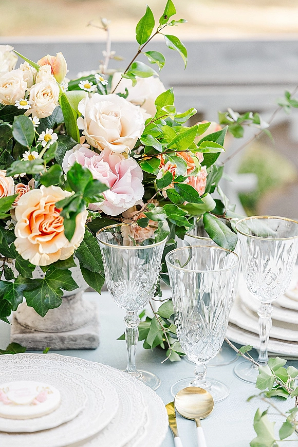 Reception tablescape with wedding table setting, peach and blush floral centerpiece, crystal goblets, gold flatware, and light blue tablecloth by railing