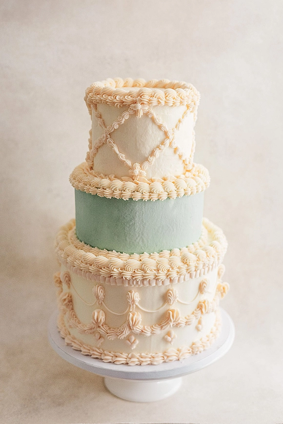 Wedding cake with three tier wedding cake design, scalloped buttercream piping and a pastel blue middle tier on a stand against a neutral backdrop