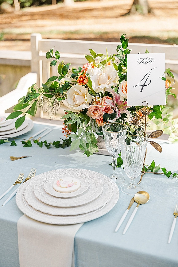 Wedding tablescape with floral centerpiece and greenery garland on a light blue tablecloth, gold flatware, crystal goblets, and garden fence backdrop