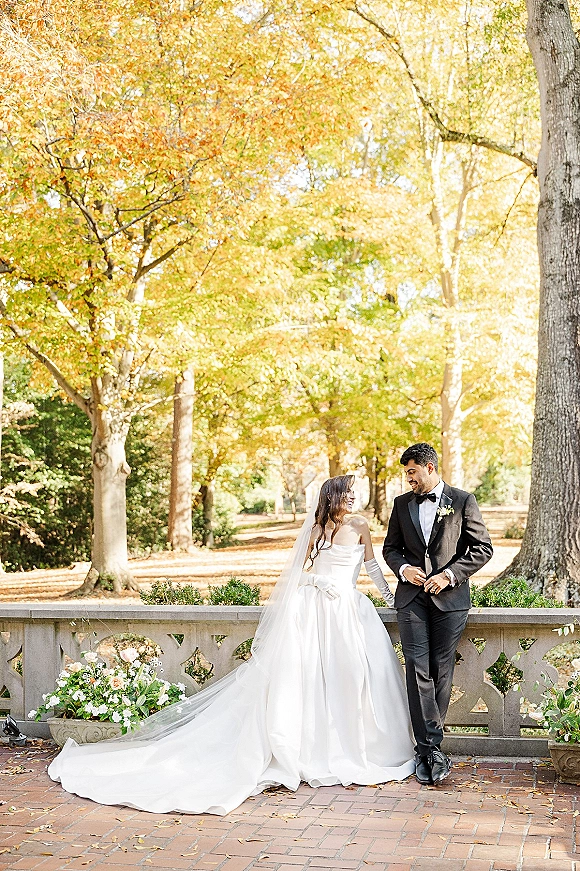Couple portrait of bride and groom laughing, leaning on a stone balustrade with autumn trees, fallen leaves, and floral arrangements behind them