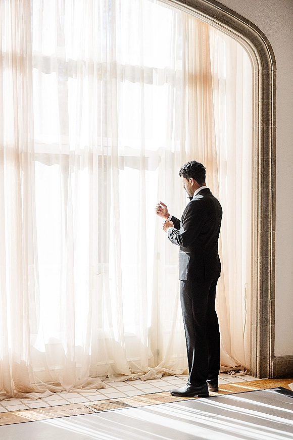 Groom portrait in a black tuxedo adjusting cufflinks by a sunlit arched window, sheer curtains behind and tiled floor below