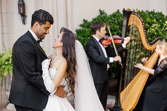 First dance as bride in veil and long gloves sways with groom in tuxedo beside a harpist and violinist on an outdoor patio