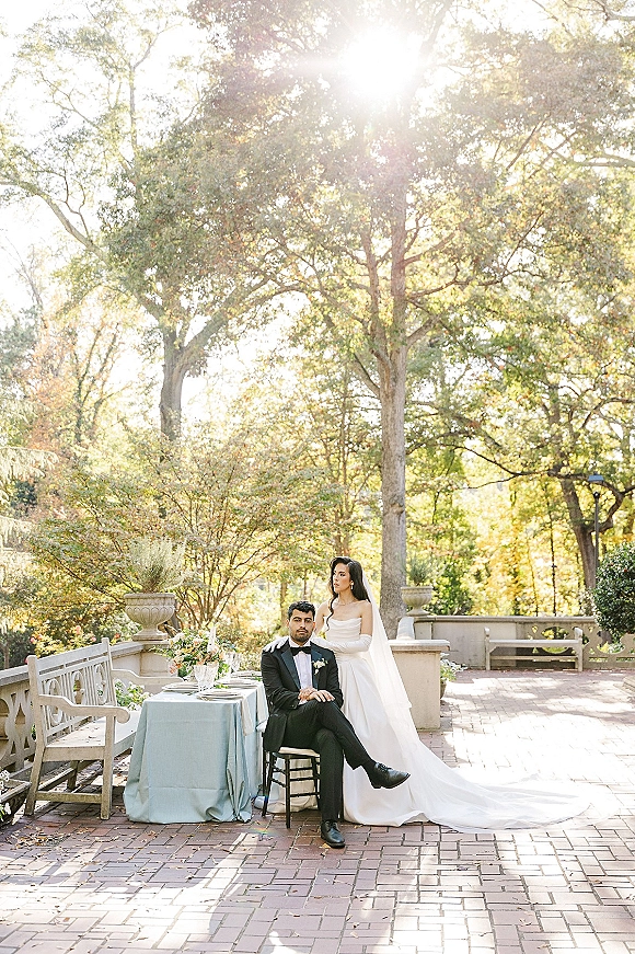 Couple portrait of bride standing behind groom seated at an outdoor reception table on a sunlit brick patio with stone planters