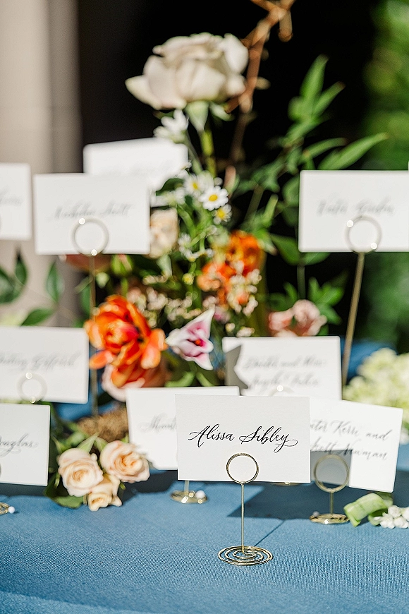 Wedding place cards with calligraphy in gold card holders on a blue linen table, accented by roses, daisies, and greenery in sunlight