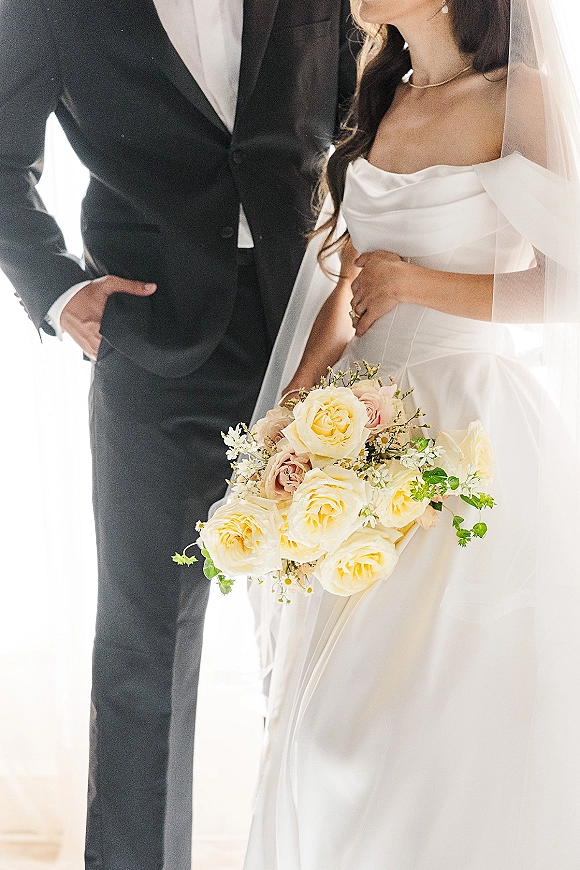 Couple portrait of bride and groom close up, bride holding bouquet of yellow roses beside groom in black tuxedo by bright window light