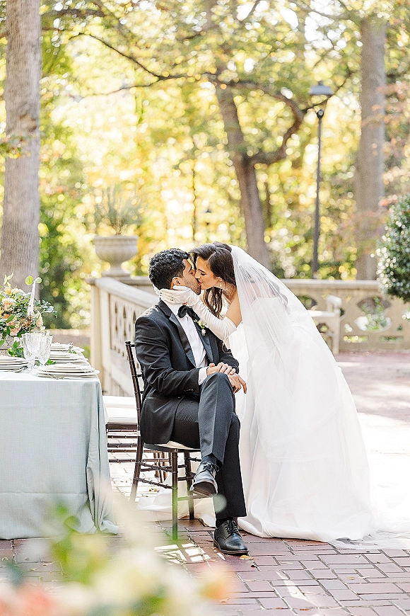 Wedding kiss portrait of bride kissing groom in black tuxedo, veil and long gloves by a reception table on a brick terrace
