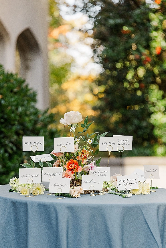 Escort card table with wedding escort cards and calligraphy place cards in gold holders, blue linen, and rose centerpiece in sunlit garden walkway