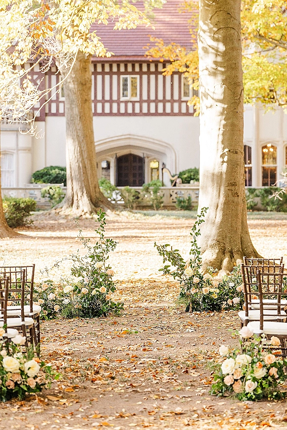 Outdoor ceremony aisle with garden wedding aisle chair rows and low rose-and-greenery markers on a leaf-covered lawn beneath large trees
