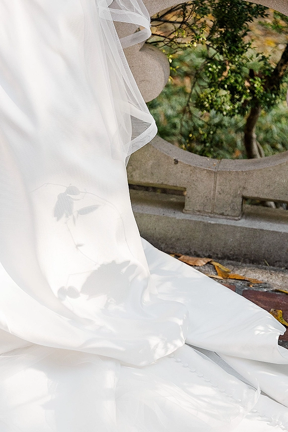Wedding dress detail with bridal veil detail, tulle veil layers draping over a clean satin train beside a stone railing in dappled sunlight