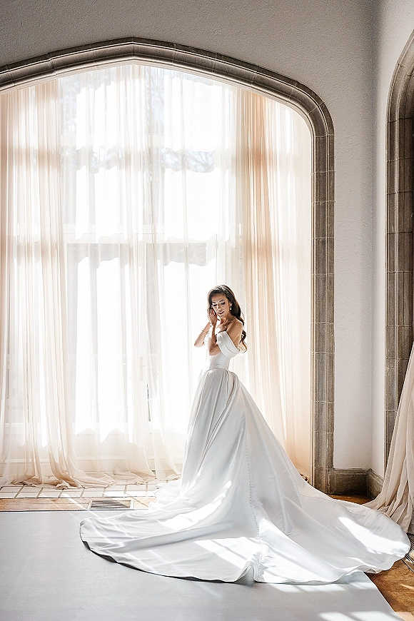 Bridal portrait of a bride in an off the shoulder wedding dress with a long train by an arched window, sunlight through sheer curtains