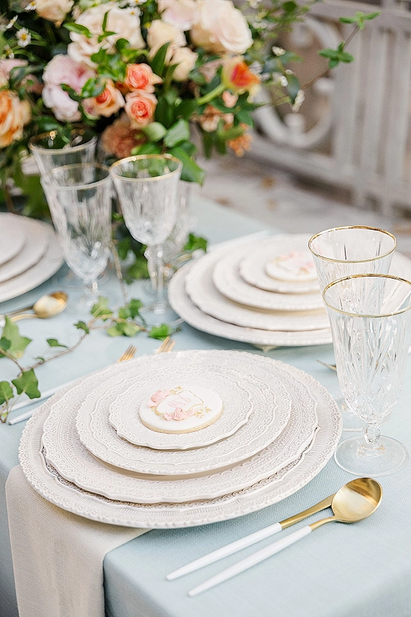 Reception tablescape with a wedding place setting of white patterned plates, gold flatware, gold-rimmed crystal glasses, and floral centerpiece on outdoor patio