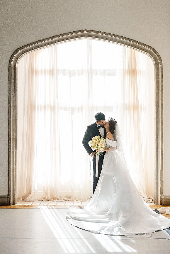 Wedding kiss portrait of bride and groom kissing by an arched window, bouquet in hand as sunlight glows through sheer curtains