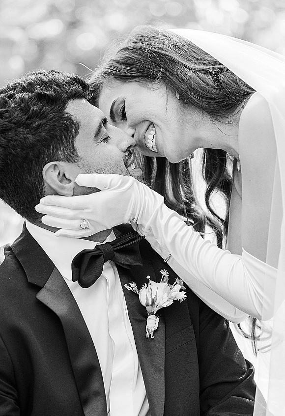 Wedding couple portrait of bride cupping groom’s face for a close-up kiss, her veil and long gloves against soft outdoor greenery bokeh lights