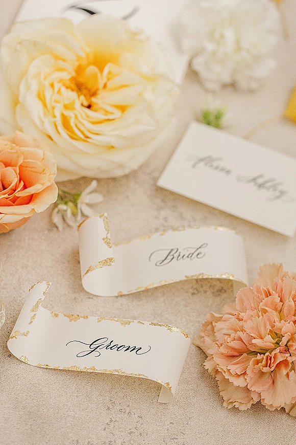 Wedding place cards with calligraphy place cards, gold leaf edging and peach florals tied with ribbon on a light stone surface flat lay