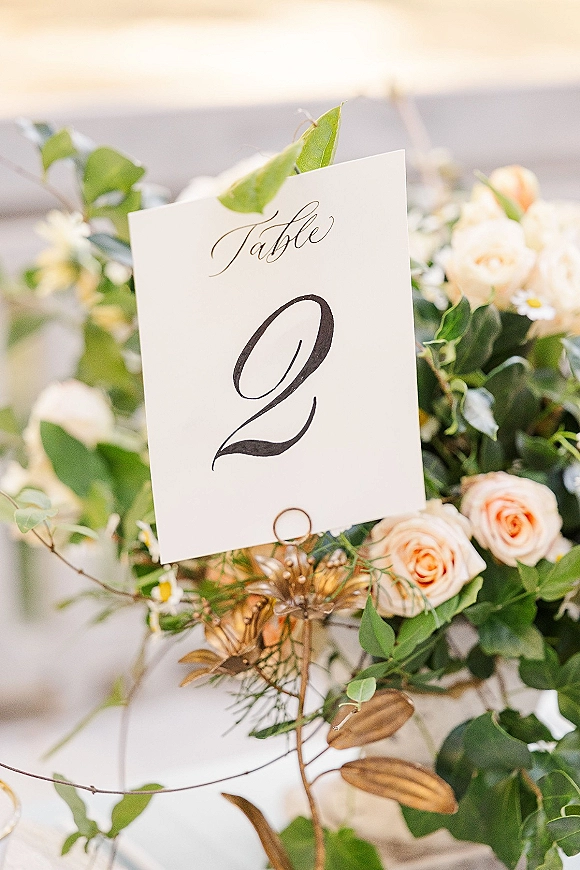 Wedding table number card with calligraphy table numbers in a gold holder beside roses and eucalyptus greenery on a softly lit table outdoors