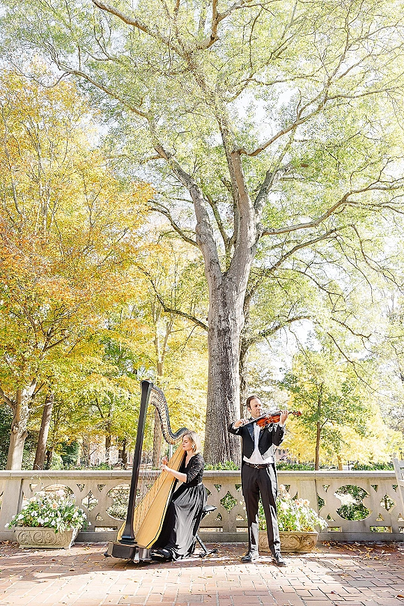 Wedding musicians playing harp and violin in formal black attire on an outdoor terrace with stone balustrade and autumn trees