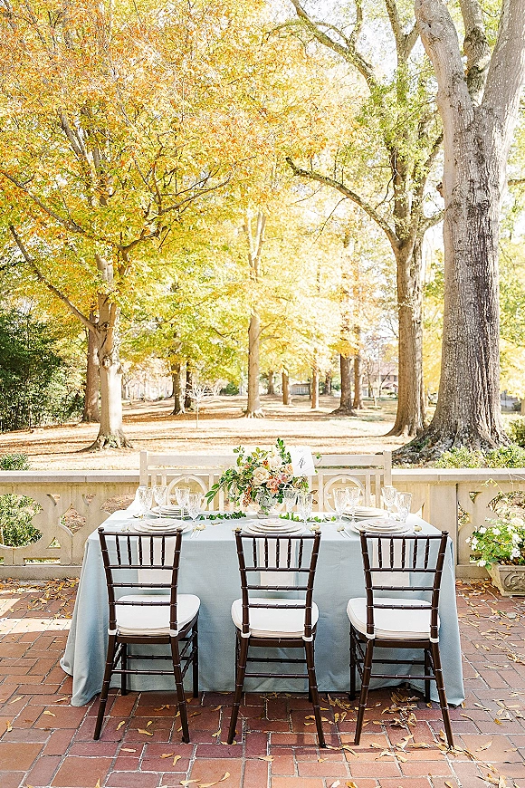 Reception tablescape with light blue tablecloth, floral centerpiece, and place cards on a brick patio beside stone balustrade and autumn trees