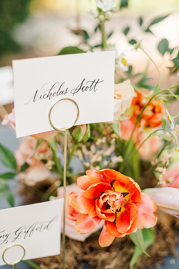 Wedding place cards with calligraphy place cards in gold holders nestled among peach roses, greenery, and an orange tulip on a bokeh reception table