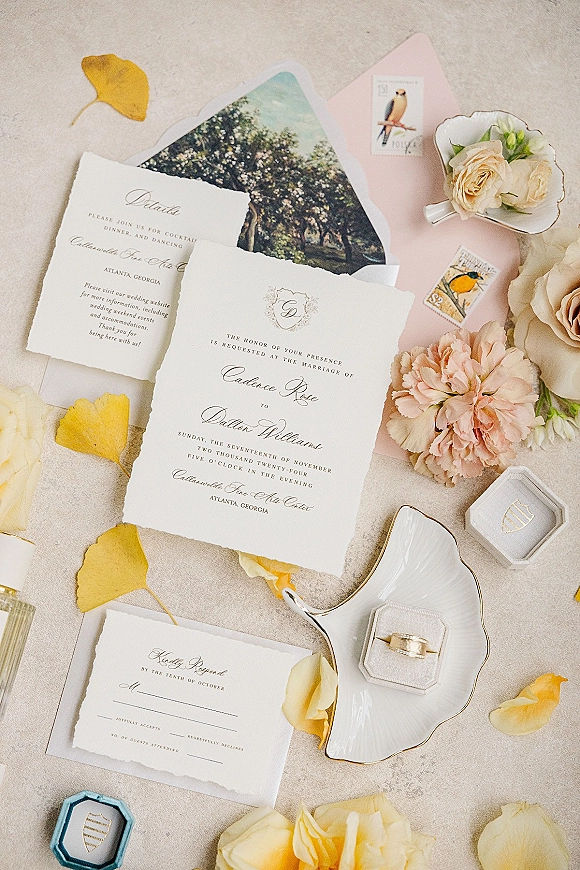 Invitation suite with deckle edge wedding invitations, envelope, and vintage stamps beside roses, peony, and rings on neutral stone surface