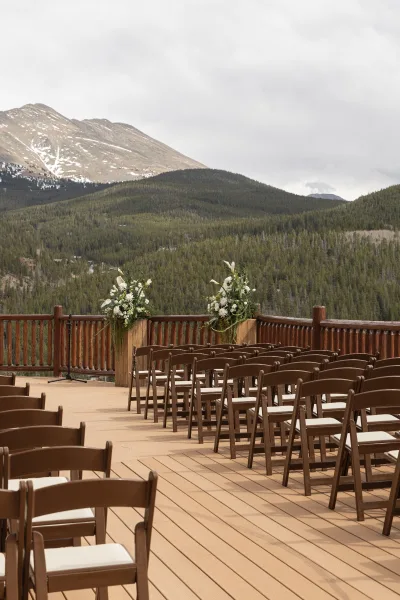 Ceremony setup with wooden folding chairs lined on a deck aisle, accented by floral pillars and greenery garland against misty mountains
