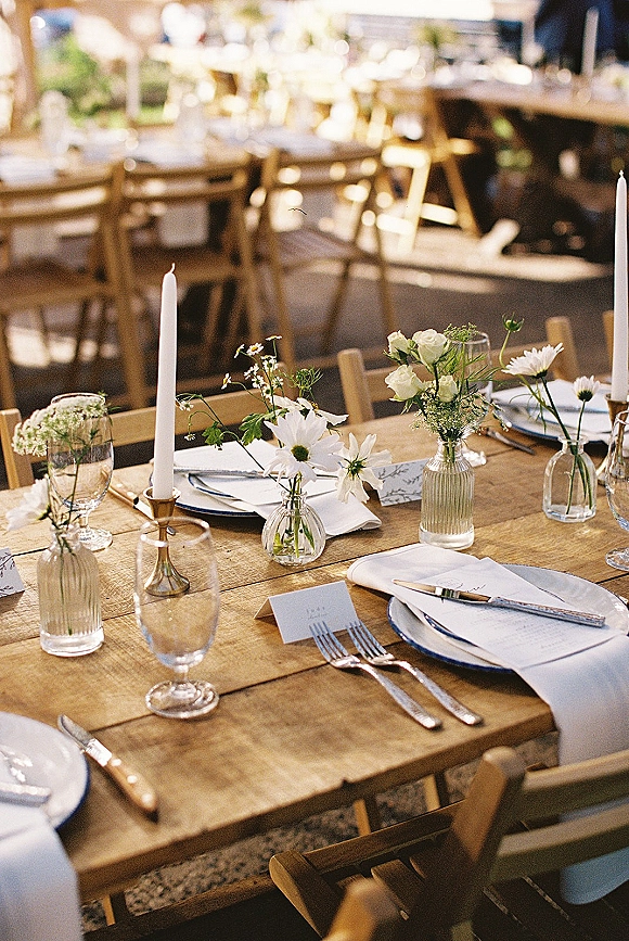 Reception tablescape with a rustic wedding tablescape on a wood farmhouse table, white taper candles, bud vases, and place cards in sunlight outdoors