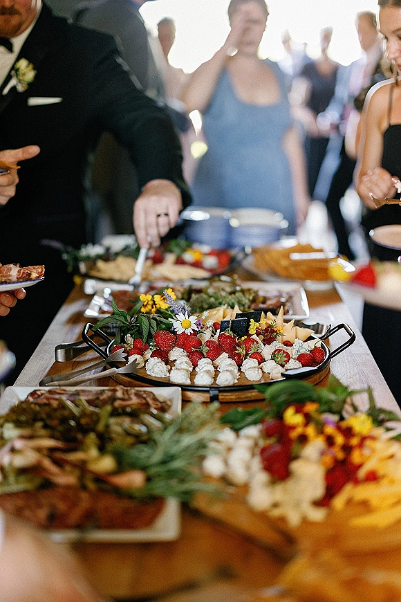 Wedding dessert table with dessert platters of strawberries, fruit, cheese, and whipped cream dollops under a tent canopy with guests nearby