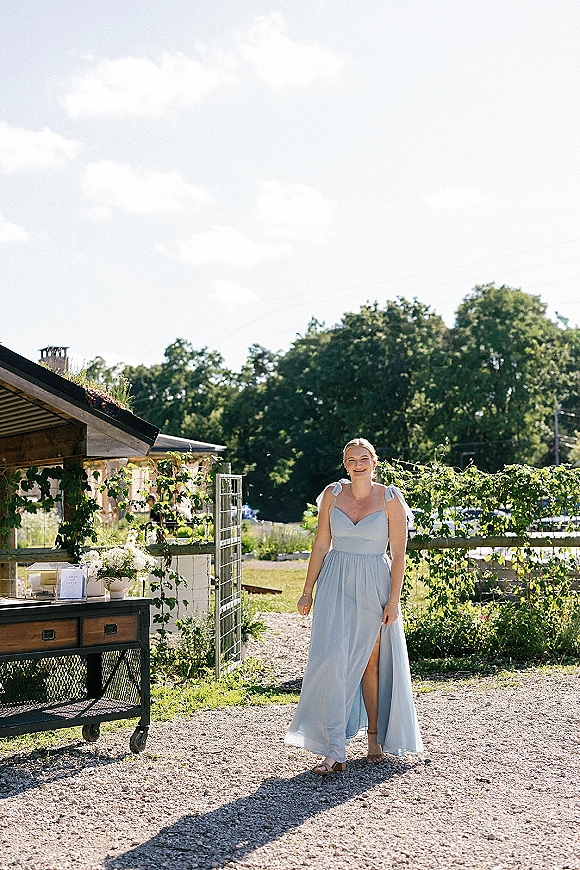 Bridesmaid portrait in a light blue bridesmaid dress with slit and bow straps, standing by a rustic vineyard welcome table in sunlight