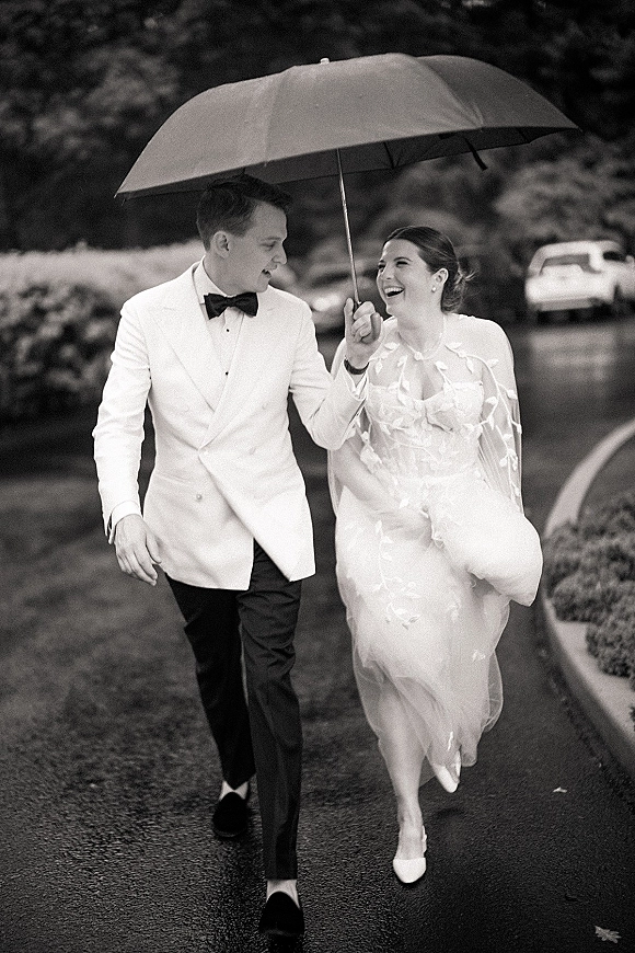 Couple portrait with wedding umbrella photo as bride and groom walk laughing on a wet road, her veil flowing beside trees and hedges