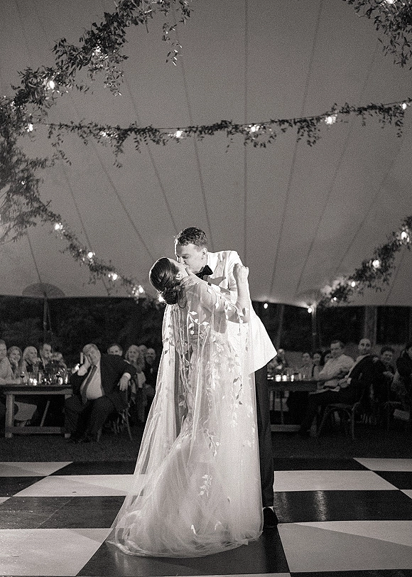 First dance as bride in wedding dress and long veil dips with groom in tuxedo under string lights and greenery in a tent reception
