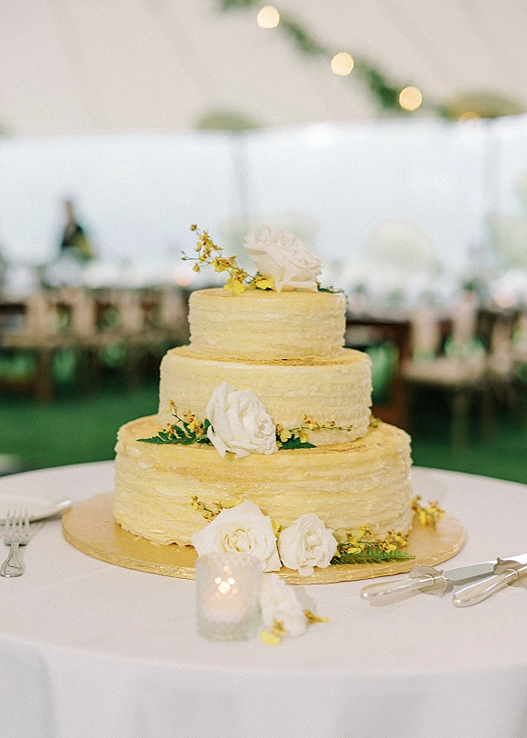 Wedding cake with textured buttercream on a cake board, topped with white roses and greenery under string lights in an outdoor tent reception