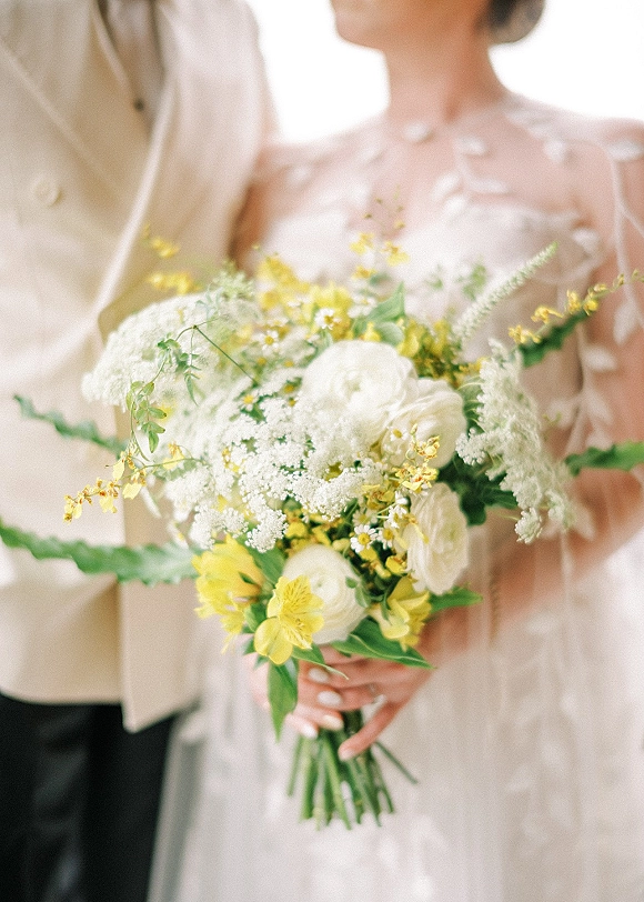 Bridal bouquet of yellow and white wedding bouquet blooms with greenery, held against a bridal gown and groom jacket in soft indoor light