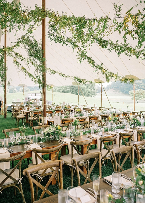 Reception tablescape in an outdoor tent reception with farm tables, greenery garlands, floral centerpieces, candles, and string lights under a sailcloth tent
